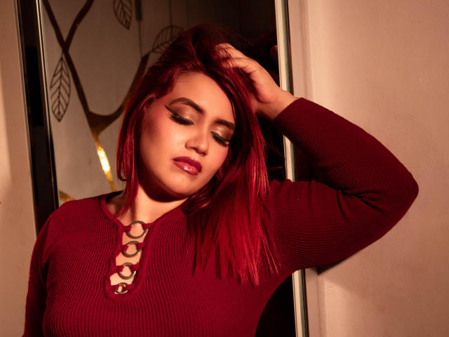 DannaScully with red hair, eyes closed, wearing a red rib-knit top with lace-up detail, hand in hair, red lipstick, warm light, sexy, soft expression, medium shot.