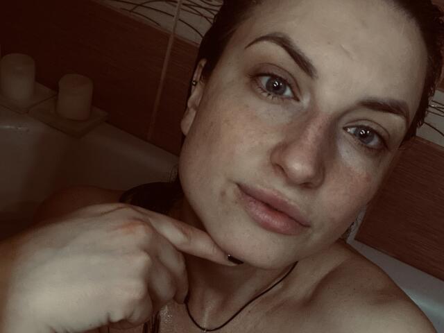 Milagres has a fair-skinned face with dark brown eyebrows, blue eyes, and wet hair, touching her chin with a finger in a dimly lit bathroom.