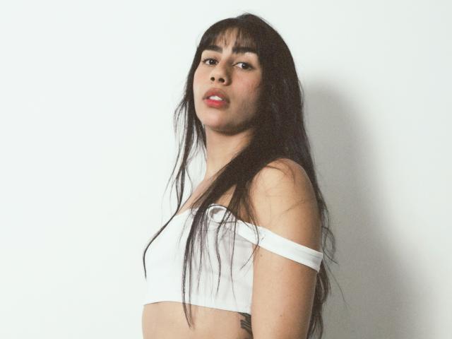 JessicaSoler with long black hair, wearing a white off-shoulder top, exposing her small tits, standing against a plain white background, looking at the camera with red lips.