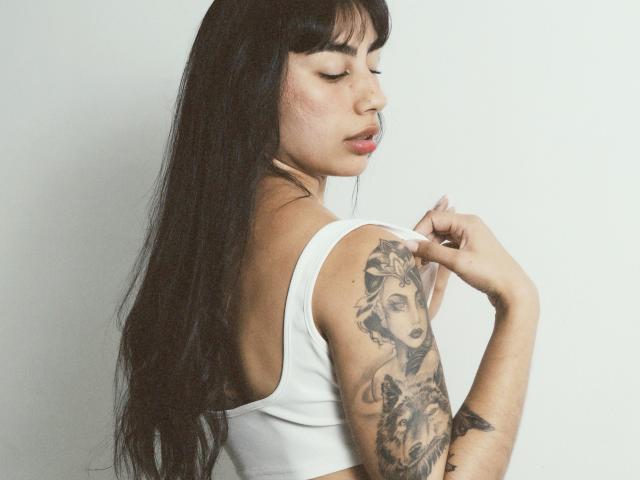 JessicaSoler with long black hair, wearing a white tank top, showing a detailed tattoo of a woman's face on her right arm, touching her shoulder.