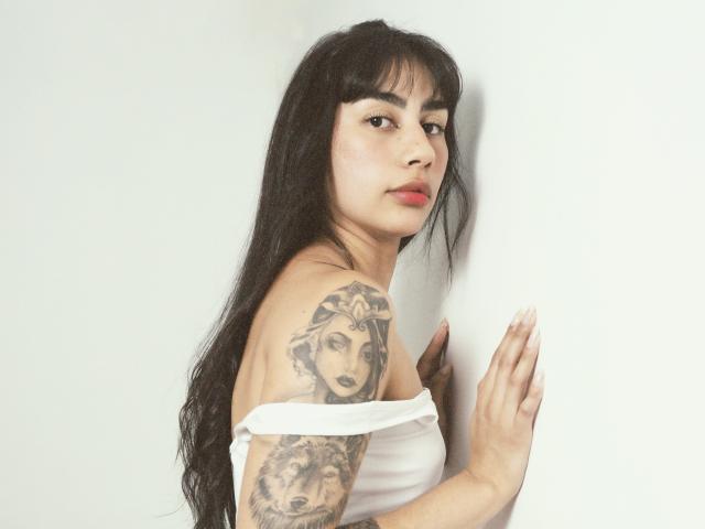 JessicaSoler with long black hair, red lipstick, white off-shoulder top, tattoo of wolf-faced woman on right arm, hand against white wall, looking over shoulder, soft light.