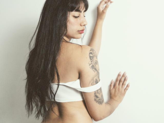 JessicaSoler with long black hair, white off-shoulder top, tattoo on right arm, back to camera, eyes closed, hand on wall, ass slightly visible, soft lighting, white background.