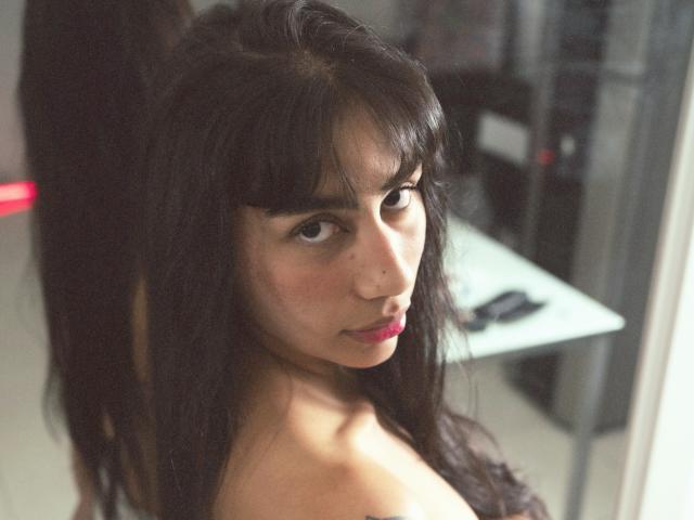 JessicaSoler with dark, wavy hair and bangs, looking over her shoulder with a slightly pouty expression, bare shoulders, and a subtle smile, in a softly lit room.