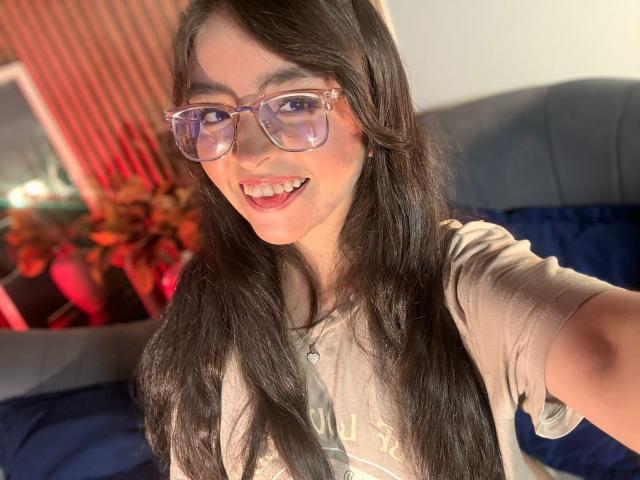 EvaMcAllister with long black hair, wearing clear glasses and a beige t-shirt, smiling with her tongue slightly out, showing her teeth. Her boobs are partially visible.