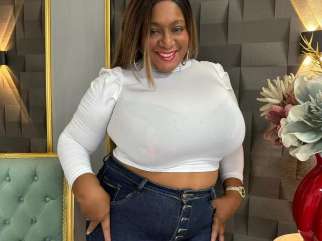 NahomiAnders with huge tits, wearing a white long-sleeve shirt and tight blue jeans, sitting on a gold chair, smiling with long brown hair, wearing a blue wristwatch.