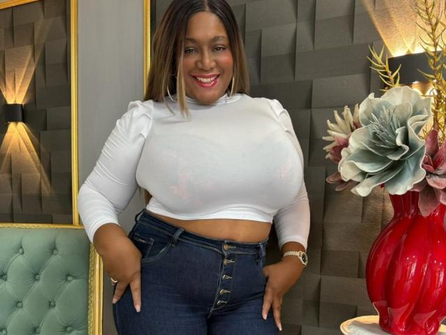 Nahomi Anders, com pele morena, cabelos longos e lisos castanhos, camisa branca de manga comprida e calças jeans azuis justas, curvada sobre uma cadeira verde, exibindo suas nádegas grandes e sorrindo..