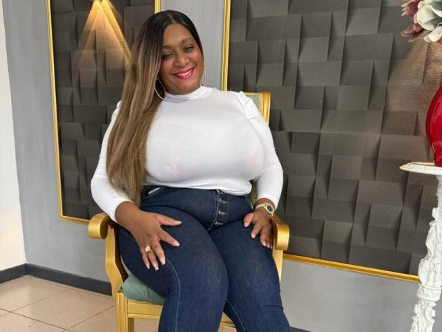 Nahomi Anders, com seios grandes e visíveis, vestindo uma blusa branca transparente, calças jeans escuras e com um sorriso. Um vaso vermelho com flores grandes à direita, arandelas douradas na parede..