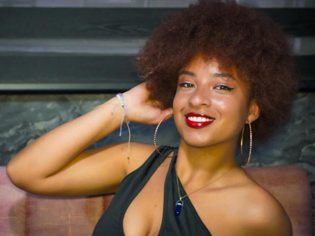 NiaZuri has a curly brown afro, red lipstick, and black halter top, smiling with her hand in her hair, wearing hoop earrings and a blue necklace.