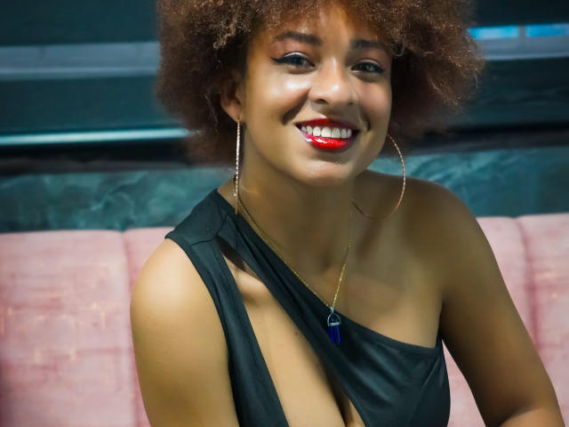 NiaZuri with curly brown hair, red lipstick, black one-shoulder dress, showing her cleavage, smiling, wearing large hoop earrings, and a blue necklace, sitting on a pink bench.