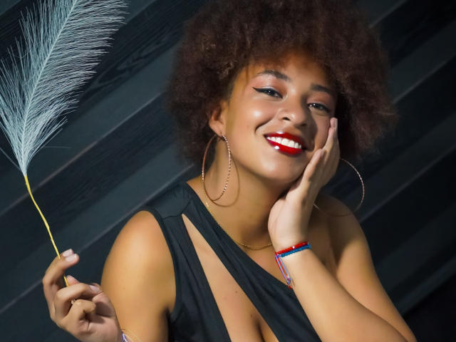 NiaZuri with curly brown hair, red lipstick, smiling, holding a feather, wearing a black dress, hoop earrings, and a colorful bracelet, touching her cheek. Boobs visible through dress.