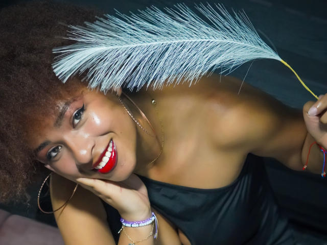 NiaZuri with curly brown hair, red lipstick, holding a white feather, wearing a black strapless dress, smiling, looking up, wearing a bracelet and hoop earrings, dark skin.