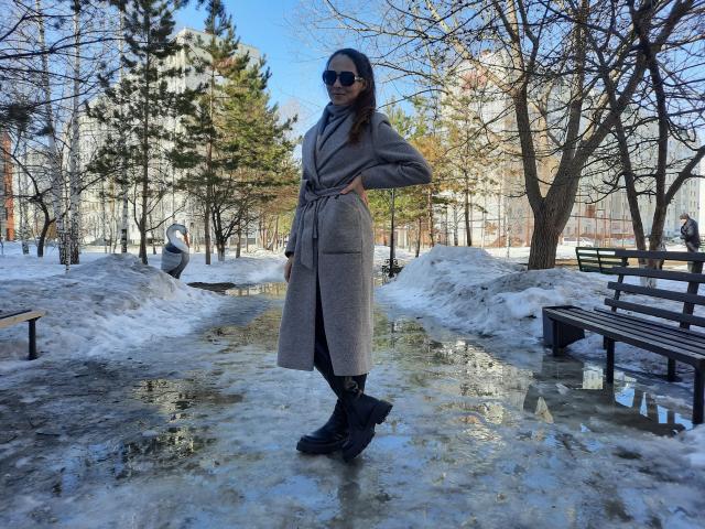 MissKinney stands in a snowy park wearing a grey coat, black boots, and sunglasses, with hands on hips, next to a puddle with a tree and bench in background.