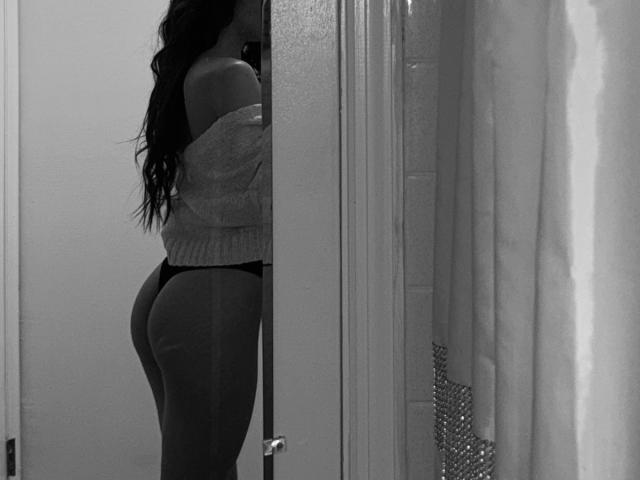 DianaFernandezz with long dark hair, wearing an off-shoulder sweater and black thong, showing her ass in a mirror selfie, black and white, reflection, door in background.