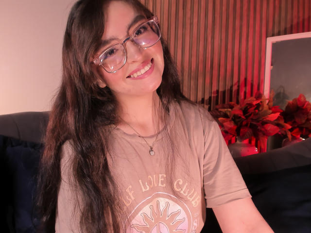 EvaMcAllister with long dark hair, glasses, smiling, wearing a beige "Love Club" t-shirt, necklace, sitting, red plant, and black chair in a warmly lit room.