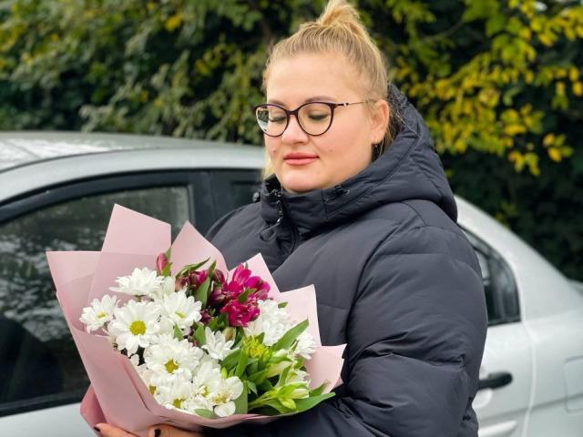 RadaRori with glasses, blonde ponytail, black puffy jacket, holding pink-wrapped bouquet of white daisies and purple flowers, standing next to silver car.