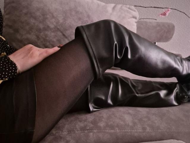 Anshante's legs in black sheer tights and shiny black leather boots, lying on a grey sofa. A hand rests on her thigh. Boobs and pussy are not visible.