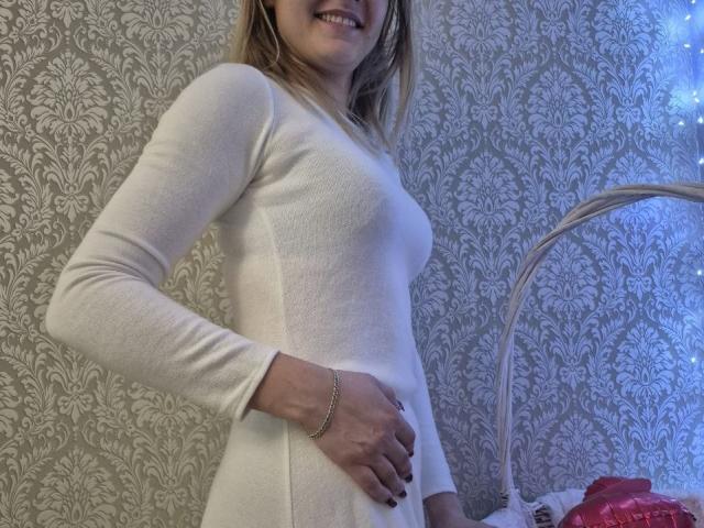 Anshante wearing a tight white long-sleeve dress, hand on her ass, smiling, blonde hair, silver bracelet, blue light on right. Her tits are slightly visible through the fabric.