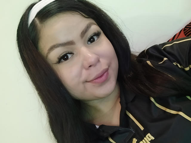 LenoreDina has long black hair, a white headband, and brown eyes. She's smiling with pink lips, wearing a black and gold shirt, showing her cute face and tits.