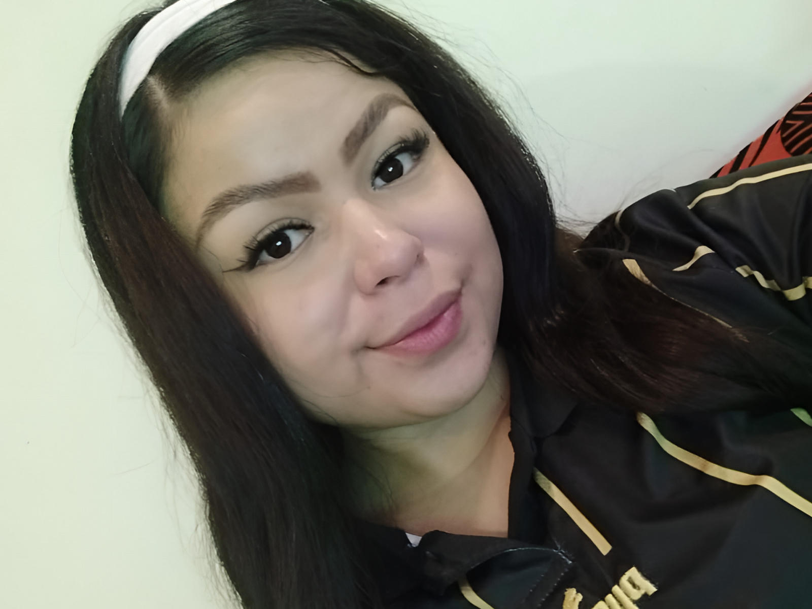 LenoreDina has long black hair, a white headband, and brown eyes. She's smiling with pink lips, wearing a black and gold shirt, showing her cute face and tits.