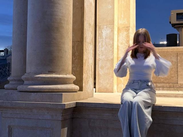 DonnettaVannuland, with hands covering face, sits on stone ledge, wearing white blouse and grey high-waisted pants, in front of large beige columns, at dusk.
