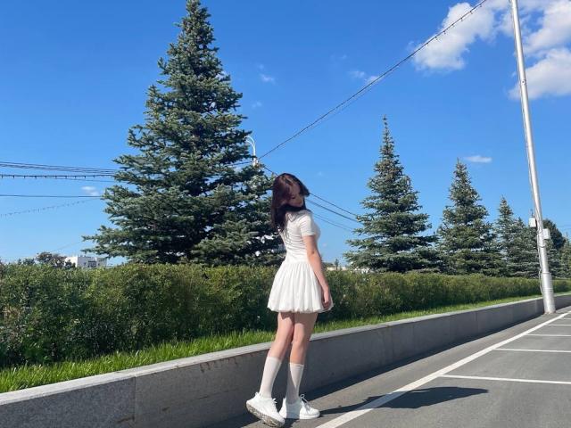 DonnettaVannuland with long brown hair, white short-sleeve dress, white knee socks, and white sneakers, standing on a sidewalk, looking back, in front of tall green trees.
