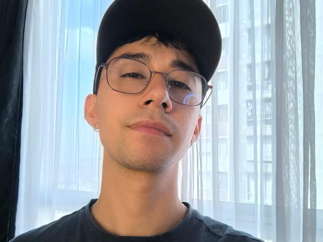 OliverHot69 com óculos, boné preto, cabelo preto curto, pele clara, sorriso discreto, pequena argola no ouvido, vestindo camisa escura, luz suave, leve sombra, expressão relaxada, ambiente interno..