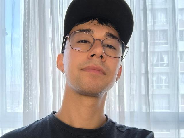 OliverHot69 es un chico atractivo que usa gafas, un gorro negro y un arete, y lleva una camisa negra, con una expresión ligeramente arrogante. Tiene un tono de piel claro y cabello corto y oscuro..