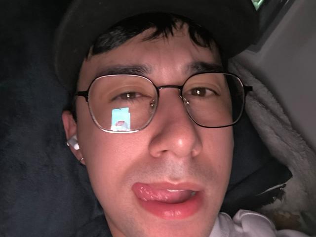 OliverHot69's face, wearing glasses and a hat, with tongue out, showing a reflection in the glass. He has a white ear piercing and a slightly sweaty forehead.