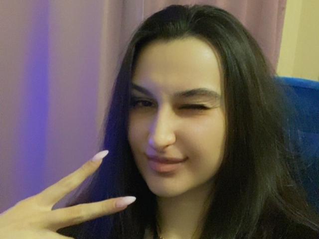 PamDeadmond has long black hair, winking with a slight smile, and is making a peace sign with her pink-manicured finger. Her cute nose is centered.
