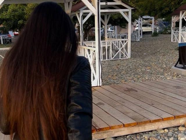 PamDeadmond with long brown hair, wearing a black jacket, stands on a wooden platform, facing white pergolas and a cobblestone area, with a fire pit to the right.