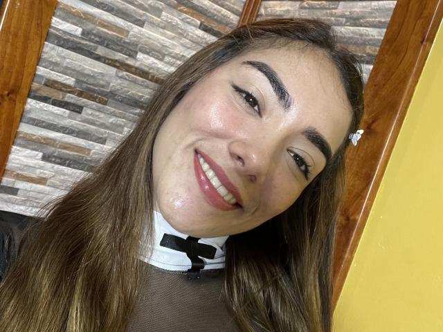 SharonKlart's smiling face with glossy skin, brown eyebrows, and straight brown hair. She's wearing a black mesh top with a white collar and black bow.