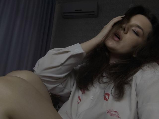 HecateCorvin, con cabello castaño largo, mejillas rosadas y maquillaje sutil, vestida con un top de encaje blanco, mostrando pequeñas tatuajes en el pecho y los brazos superiores, y con lápiz labial rojo..