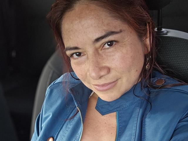 La giacca blu di AlessiaRed è aperta, rivelando il suo décolleté con un reggiseno a fiori. Indossa la cintura di sicurezza, con la lingua fuori, e ha i capelli di un castano rossiccio. Le sue labbra sono leggermente socchiuse..
