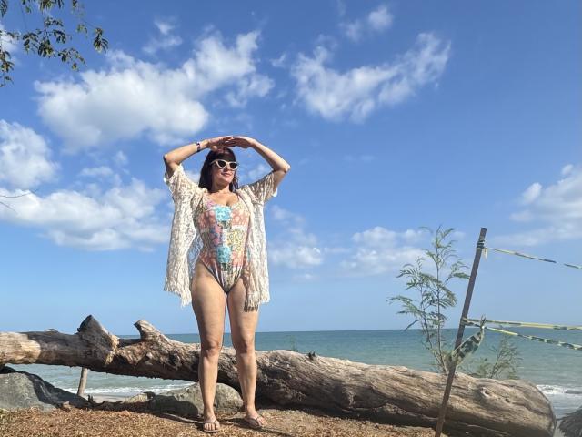 JaneRoa stands on a grassy, leafy spot with a large, fallen tree. She's wearing a floral, sheer top and a thong, showing her bare ass and small, perky tits.