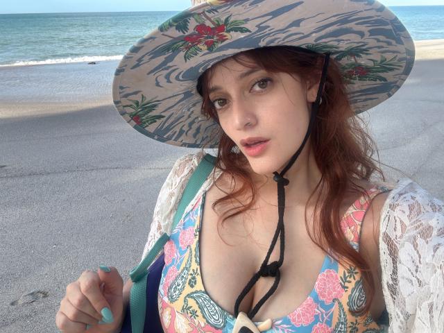 JaneRoa standing on beach, arms above head, wearing a floral one-piece, sheer white cover-up, sunglasses, and sandals, with a large driftwood log in front. Clear blue sky.