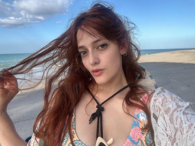 JaneRoa with red hair, wearing a floral bikini top, lace shirt, and sun hat, has a nose ring and black earbuds, showing her tits and a cute, relaxed face.