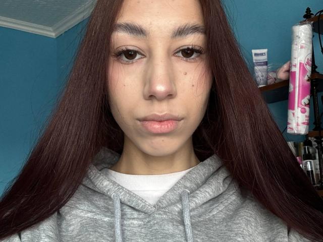 LaritaGlawson with long brown hair, grey hoodie, and serious expression, showing small nose and slightly pouty lips, brown eyes, and subtle facial freckles.