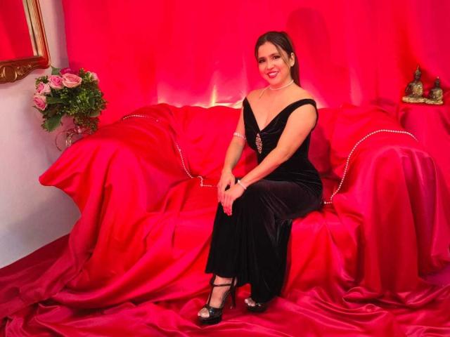 HeidyWells sits on a red satin-covered sofa, wearing a black dress with a deep neckline, black high heels, and a red necklace, smiling with pink lipstick.
