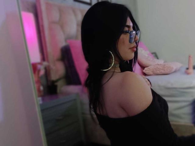IsabellaaRossi with black hair, glasses, large hoop earrings, black off-shoulder top, showing her bare shoulders and back, sitting in a bedroom with a pink headboard.