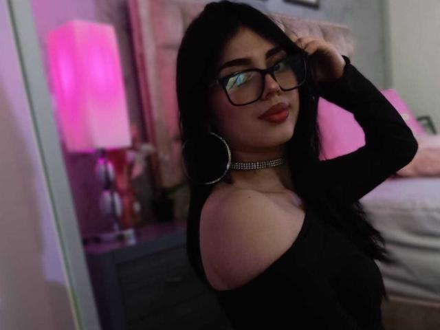 IsabellaaRossi with long black hair, glasses, and a choker, wearing an off-shoulder black top, showing her bare shoulder and smiling with red lipstick.