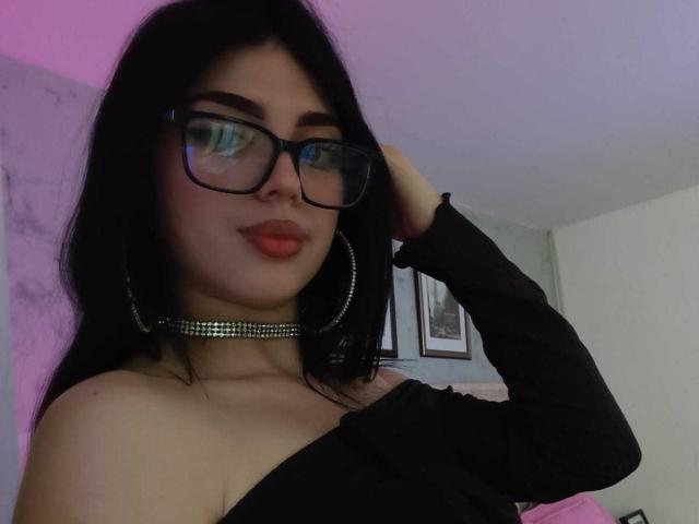 Isabella Rossi, con capelli neri, occhiali, rossetto rosso, top nero con una spalla scoperta, una collana scintillante, una mano tra i capelli, orecchini a cerchio e una luce rosa-viola..