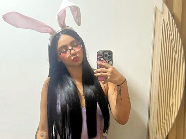 ChloeMoretti with long black hair, pink glasses, and white bunny ears, taking a mirror selfie, wearing a pink top, showing a tattoo on her right arm, and a bracelet.