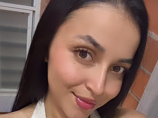 HikariChloe hat ein süßes Gesicht mit dunkelbraunen Augen, langen schwarzen Haaren und einem leichten Lächeln. Sie trägt rosafarbenen Lippenstift und ein weißes Top mit Halter..