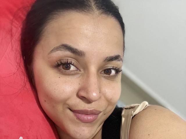 HikariChloe has a cute smile, brown eyes, and a small nose ring. She's lying on a red pillow with black and beige straps visible on her shoulder.