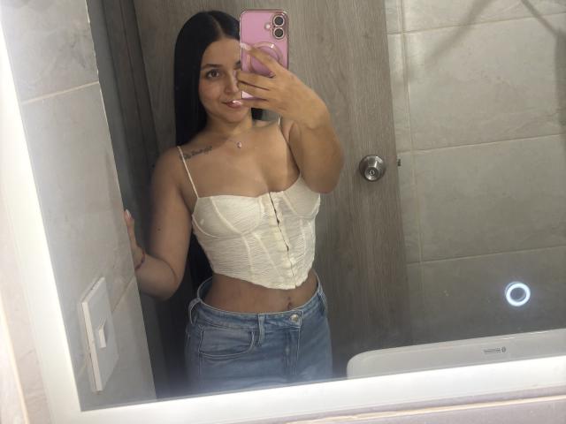 HikariChloe with long black hair, tan skin, and small tattoos, wearing a white lace corset and blue jeans, taking a mirror selfie with a pink phone.