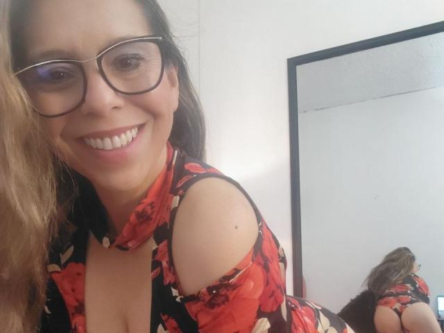 VickyVegas with glasses, smiling, red floral dress, off-shoulder, showing cleavage, mirror reflection, brown hair, white walls, black-framed mirror, butt visible in reflection, sexy.