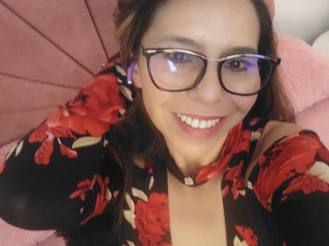 VickyVegas with glasses, smiling, wearing a black floral dress, red flowers, holding purple earbuds, pink couch, brown hair, light skin, cute, happy, selfie, medium shot.