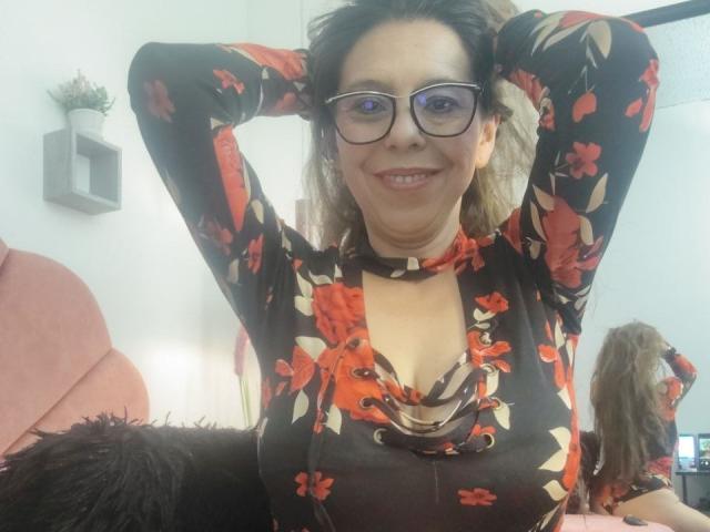 VickyVegas with glasses, smiling, hands in hair, wearing a black floral top with red and orange flowers, showing her tits, standing in a room, reflection in mirror.
