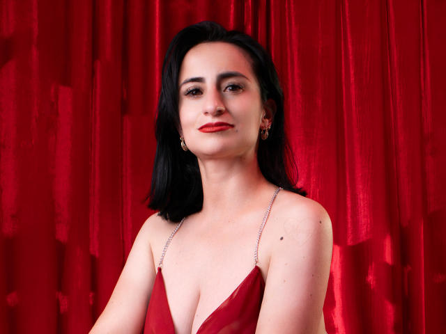 BrendaCantorel with red lipstick, black hair, and a red dress with thin straps, showing her tits against a red curtain, looking slightly to the side with a subtle smile.