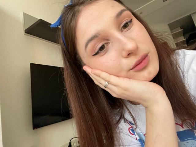 VikVikLove with brown hair, blue eyes, and winged eyeliner, resting her chin on her hand, wearing a white shirt with a blue and red pattern. She's wearing a ring.
